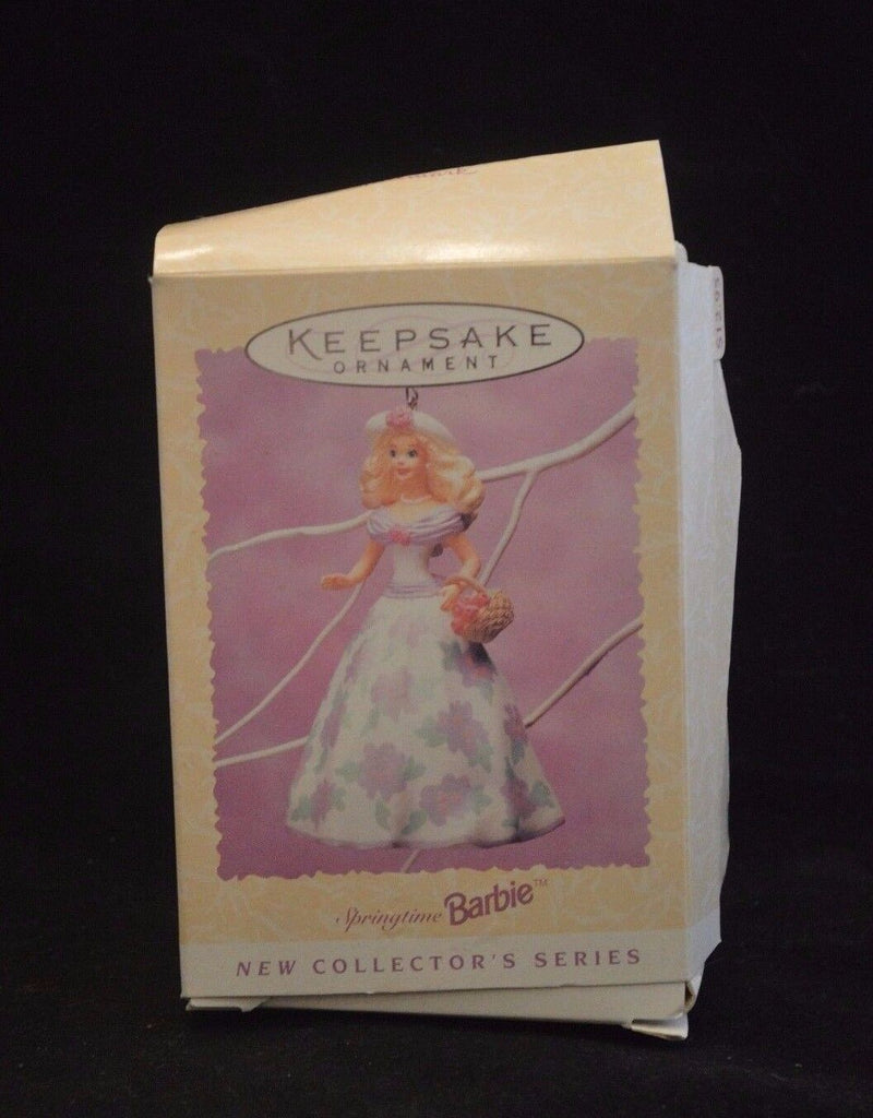 A vintage box showingcasing a Barbie springtime ornament. Barbie is set against a pink background and is holding a basket of flowers in her right hand. She is wearing a long, white floral dress with pink and green flowers, a white sunhat with a pink flower on the front, and a pearl necklace. The box's text says, "KEEPSAKE ORNAMENT - Barbie - New Collector's Series".