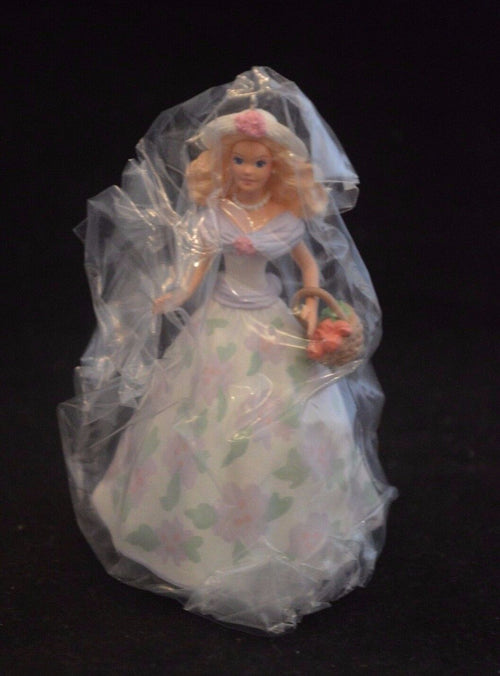 A Barbie ornament wearing a long, white floral dress with pink and green flowers, a white sunhat with a pink flower on the front, and a pearl necklace. Barbie is holding a basket of roses in her right hand. She has blonde, curly hair.